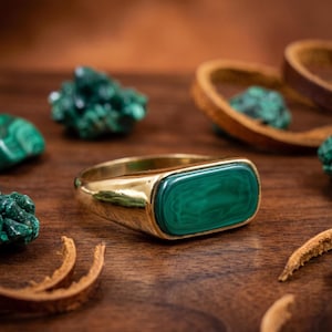 May include: A gold-toned ring featuring a smooth, rectangular, horizontally oriented malachite inlay with distinct dark green banding. The band is polished and solid, showcasing a clean, minimalist design that highlights the natural stone.