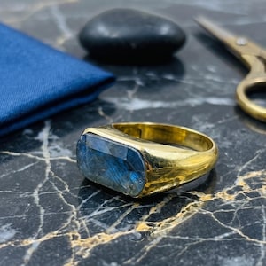 May include: A gold ring featuring a rectangular, faceted labradorite gemstone with blue iridescence. The band is smooth and polished, showcasing a wide, solid metal construction that tapers slightly toward the back of the ring setting.