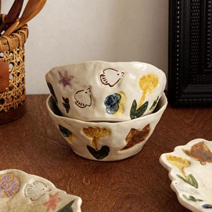 以下が含まれることがあります： 重ねられた2つのオフホワイトのセラミックボウル。カラフルな花と鳥のデザインで装飾されています。ボウルは質感のある有機的な形をしています。お揃いのプレートが前景に見えます。木製の食器が背景の織りバスケットに入っています。