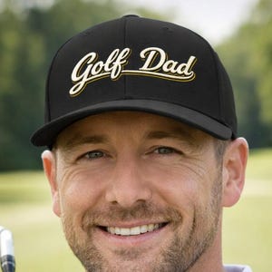 May include: Black baseball cap with the text "Golf Dad" in gold script. The cap has a curved brim and is worn by a person holding a golf club. The person is smiling and wearing a light blue collared shirt.