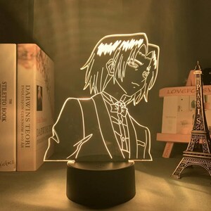 May include: A glowing anime-style character lamp. The lamp features a detailed outline of a person in a suit and bow tie, illuminated with a soft white light. The lamp sits on a black base, with books and a miniature Eiffel Tower in the background.