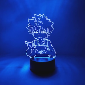 May include: A blue LED lamp featuring a white outline of an anime character eating from a bowl with chopsticks. The lamp has a black base with a power button and emits a bright blue glow against a dark blue background.