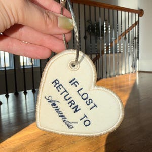 Puede incluir: Una etiqueta para equipaje en forma de corazón crema con el texto bordado en azul marino "IF LOST RETURN TO Amanda". Tiene un lazo de metal plateado para sujetarla.