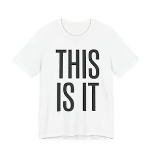 Puede incluir: Camiseta blanca con la frase "THIS IS IT" en letras negras y en negrita. La camiseta tiene mangas cortas y cuello redondo. El texto está centrado en la parte delantera de la camiseta.