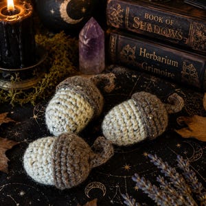 May include: Three crocheted acorns featuring cream-colored bodies and textured brown caps with small curled stems. Each acorn is crafted from yarn with a visible stitch pattern, showcasing a two-tone design that mimics the appearance of natural nuts.
