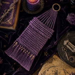 May include: A purple crocheted wall hanging featuring a circular gold-toned metal ring at the top, a rectangular woven body, and a bottom fringe accented with small, pointed gold-toned metal spikes attached to the base of the woven section.