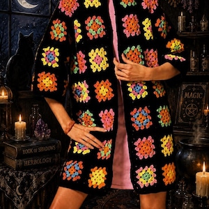 May include: A black open-front cardigan featuring a repeating pattern of colorful crochet granny squares in shades of pink, orange, yellow, green, and blue. The garment has three-quarter length sleeves and a straight hemline, worn over a light pink base.