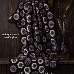 Pode incluir: Um cobertor Midnight Rosette Motif Afghan, feito de crochê com um padrão floral em preto, roxo e creme. O cobertor está sobre uma superfície de madeira. O texto "The Christmas Witch shop" e "Crochet Patterns & Spooky Vibes" são visíveis.