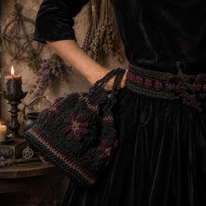 May include: A black crochet bag with a floral pattern in burgundy and tan is held by a person wearing a black velvet dress. A matching crochet belt with burgundy accents is also visible. The scene is set with candles and dried herbs.