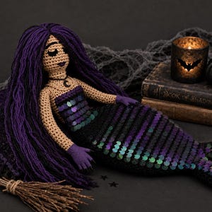 May include: A handmade crochet mermaid doll with long purple hair, a black and purple sequined tail, and a black top. A small broom, a candle holder with a bat silhouette, and a small pumpkin are also in the image, suggesting a Halloween theme.