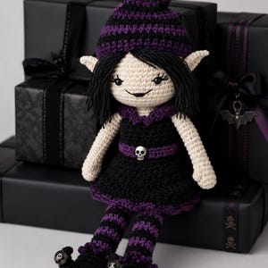 May include: A handmade crochet doll with a gothic theme. The doll has a cream-colored face, black hair, and is wearing a black dress with purple accents. The doll's hat and striped socks are also purple and black. The doll is sitting on a stack of black gift boxes.