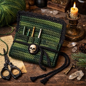 Puede incluir: Un estuche de ganchillo verde y negro con un cierre de calavera. El estuche contiene varios ganchillos. El estuche está rodeado de una madeja de lana, tijeras, una vela y cristales.