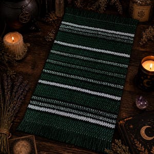 May include: A green and white striped rectangular textile with a fringed edge. The textile is placed on a dark wooden surface, surrounded by candles, books, and dried herbs. The overall aesthetic suggests a mystical or witchy theme.