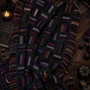 May include: A handcrafted, woven blanket with a geometric pattern in shades of purple, green, and orange. The blanket has a fringed edge and is displayed on a dark wooden surface. The blanket is approximately 152 cm by 127 cm.
