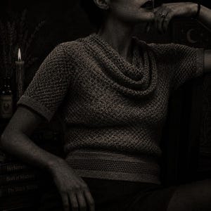 May include: A woman wearing a short-sleeved, textured knit sweater with a cowl neck. The sweater is a muted gray color. The image has a vintage, gothic aesthetic, with a candle, books, and a potion bottle in the background.