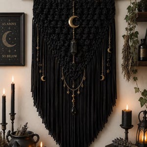 May include: Black macrame wall hanging with gold moon and star accents. The wall decor features a central crystal pendant and dangling chains. A framed print reads "As Above, So Below."