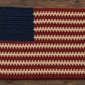 May include: A rectangular crochet piece featuring the design of the American flag. The left section is a solid dark blue block, while the remaining area consists of alternating horizontal stripes in deep red and off-white yarn textures.