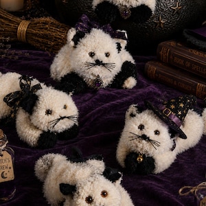 May include: A collection of fluffy white and black cat toys, some adorned with bows and witch hats, are displayed on a purple velvet cloth. The image includes the text "The Christmas Witch shop Crochet Patterns & Spooky Vibes".