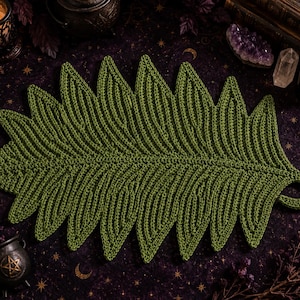 May include: A dark olive green crochet mat shaped like a large fern leaf. The textile features a central spine with symmetrical, ribbed, pointed segments extending outward, and a small circular loop at the base for hanging or storage purposes.