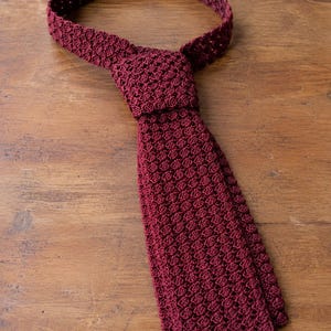 May include: A deep burgundy necktie crafted with a dense, textured crochet knit pattern. The tie features a consistent, repeating stitch design throughout its length, including the knot and the wide blade, showcasing a uniform dark red color.