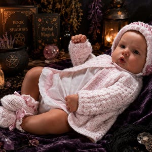 May include: A baby doll wearing a light pink and white crocheted cardigan, a matching bonnet with pink ribbon trim, and white crocheted booties with pink ribbon bows. The doll is dressed in a white onesie and has a realistic facial expression.