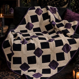May include: A cozy, handmade blanket with a geometric pattern. The blanket features a repeating design of black squares and purple diamond shapes against a neutral beige background. The blanket is draped over a black couch, creating a warm and inviting atmosphere.