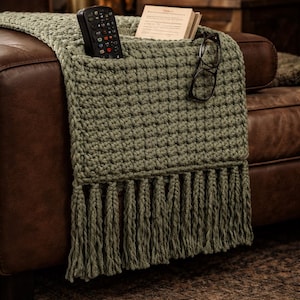 May include: Olive green crocheted throw blanket with a pocket and fringe detail. A remote control, glasses, and a small book rest in the pocket. The blanket is draped over a brown leather couch.
