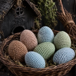 Puede incluir: Una rústica cesta de mimbre llena de siete huevos de ganchillo en tonos marrón, rosa, azul y verde. Los huevos están colocados entre musgo seco. Una pequeña casa de madera y una escoba en el fondo, creando un tema artesanal.