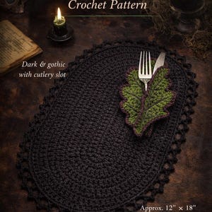 May include: Black oval crochet placemat with a scalloped edge, designed for a witch's table. Includes a leaf-shaped cutlery holder with a fork and knife. The text reads "Witch's Table Oval Placemat Crochet Pattern". The placemat measures approximately 12" x 18".