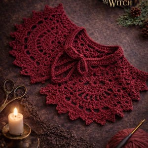 Puede incluir: Un cuello de ganchillo burdeos con un borde festoneado y un cierre de lazo. El cuello se muestra sobre una superficie oscura y texturizada con una vela encendida, tijeras y el texto "The Christmas Witch".
