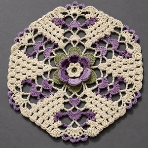 May include: A cream-colored, crocheted doily with a floral design. The doily features a central flower with purple petals and green leaves, surrounded by purple and cream-colored accents. The edges are scalloped.