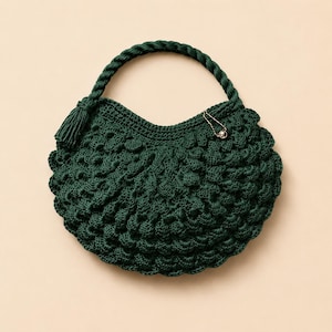 May include: A dark forest green crochet handbag featuring a textured bobble stitch pattern throughout the body. The bag has a thick, twisted rope handle and a small decorative gold-toned safety pin accent attached to the upper edge.