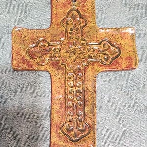 May include: A ceramic cross wall hanging featuring a mottled yellow and reddish-brown glaze. The surface displays a raised, embossed scrollwork pattern centered on the cross, with a small circular hole at the top for hanging purposes.