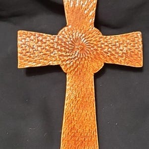May include: A copper-toned wall cross featuring a textured surface with a repeating pattern of small, elongated indentations. The center of the cross displays a circular motif with radiating lines, and a small hole is visible at the top for hanging.