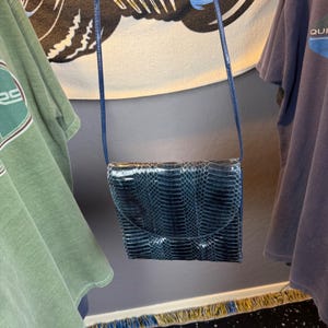May include: A blue snakeskin-patterned crossbody bag with a flap closure. The bag has a thin, matching blue strap. The bag is rectangular with rounded edges and a glossy finish. The bag is hanging in front of a gray wall.