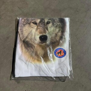 May include: A white t-shirt featuring a detailed close-up of a wolf's face. The shirt is packaged in a clear plastic bag. A circular patch with a wolf silhouette and the words "Defenders of Wildlife" is visible.