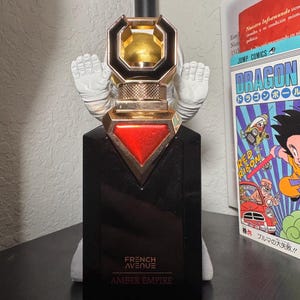 May include: A black perfume bottle with gold and red accents, featuring the text "FRENCH AVENUE AMBER EMPIRE." The bottle has a unique cap design and is set against a white background. A Japanese comic book is visible in the background.