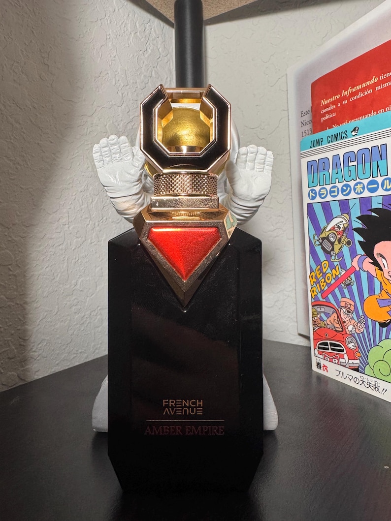 May include: A black perfume bottle with gold and red accents, featuring the text "FRENCH AVENUE AMBER EMPIRE." The bottle has a unique cap design and is set against a white background. A Japanese comic book is visible in the background.