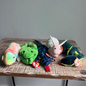 May include: A collection of five plush animal toys including a multi-colored chameleon, a bright green frog, a blue and black spotted lizard, a light pink snail with a white shell, and a dark green snake, each featuring a red heart-shaped Ty tag.