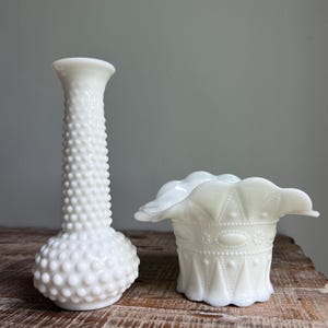 May include: A tall white milk glass vase with a hobnail texture and a narrow neck alongside a shorter white milk glass bowl featuring a ruffled edge and a geometric pattern of raised dots, lines, and oval shapes around the exterior body.