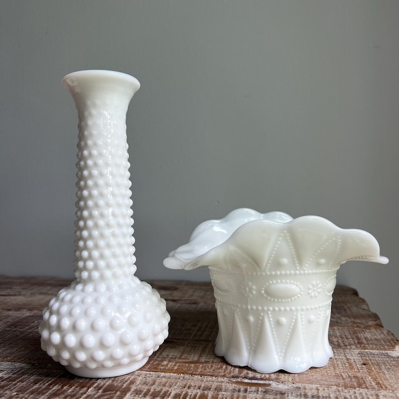 May include: A tall white milk glass vase with a hobnail texture and a narrow neck alongside a shorter white milk glass bowl featuring a ruffled edge and a geometric pattern of raised dots, lines, and oval shapes around the exterior body.
