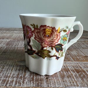 May include: A white ceramic mug featuring a scalloped edge and a handle on the right side. The exterior is decorated with a floral pattern in shades of pink, yellow, light blue, and dark green, with dark brown stems and leaves.