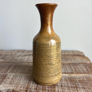 May include: A ceramic vase with a textured, horizontal striped pattern in shades of brown and tan. The vase has a narrow neck and a wider base. The top of the vase is a darker brown colour. The vase is on a wooden surface.