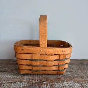 May include: A rectangular woven wood basket featuring a single upright handle. The basket is constructed from light brown horizontal wood splints interwoven with darker gray accent strips. The rim and handle are made of smooth, light-toned wood.