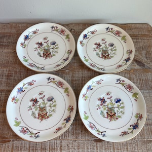 May include: Four white ceramic saucers with a central floral motif in a square planter, surrounded by a delicate vine pattern with blue, red, and green accents. Each saucer features a thin gold rim along the outer edge.