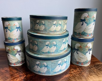 May include: A collection of vintage tin containers with lids. The containers are cylindrical and oval, featuring a painted design of white geese with blue bows against a light blue background. The lids are dark blue.