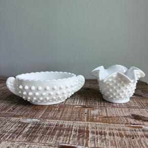 May include: A white milk glass sugar bowl with two side handles and a hobnail texture, paired with a matching white milk glass creamer featuring a ruffled rim and a hobnail pattern. Both pieces have a distinct raised dot surface design.