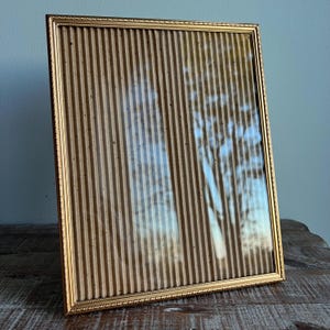 May include: A rectangular picture frame featuring a thin, metallic gold border with a decorative beaded edge. The interior of the frame contains a textured, light brown corrugated paper insert that creates a vertical ribbed pattern.
