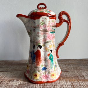 Op de afbeelding: Een porseleinen koffiepot met een handgeschilderde scène van figuren in traditionele kleding in een landschap met pagodes en kersenbloesems. De pot heeft een witte basis, een rode rand, een rood deksel en een gebogen rood handvat.