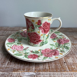 May include: A white porcelain teacup and matching saucer set featuring a detailed botanical print of deep pink roses, green leaves, and small yellow flowers. Both pieces are finished with a thin gold rim and signed by the artist Suzanne Clee.